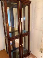 Front and side view of tall wooden curio cabinet with glass doors and gold hardware, showing interior shelving and lighting cords