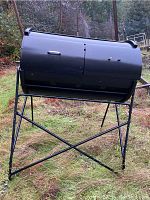 Full front view of the black metal compost tumbler mounted on stand outdoors. Shows two compartments and leg frame.