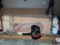 Husqvarna chainsaw placed on a wooden platform with plywood and drywall sheets in background alongside coiled rope and plastic bucket.