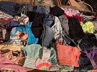 Collection of assorted little girls clothing including tops, pants, dresses, pajamas, and graphic t-shirts laid out together