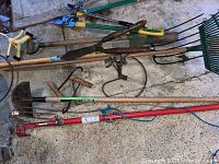 Photo showing various garden tools including shovels, rakes, hoes, pitchfork, limb cutter, and red and green handled pruning tools lying on the floor.