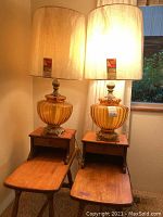 Two oak-farm style wooden side tables with one drawer each, topped with matching amber glass lamps with brass-tone bases and white shades. Positioned side by side in room corner near window.