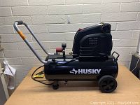 Side view of the black Husky air compressor with handle and wheels on a wooden surface against a brick wall background.