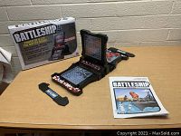 Shows the electronic Battleship game set opened with playing pieces, consoles, manual, and box visible on the table.