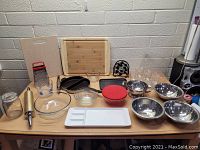 Photo of assorted kitchen items on table against gray brick wall, showing cutting boards, glass and metal bowls, measuring cup, grater, baking tray, storage container, and other utensils.