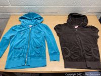 Two Juicy Girls zip up tops laid flat on a table, showing front view including colors, pocket styles, and zipper details.