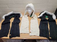 Front view of three knit hooded scarves laid out flat, two black and one white with faux fur trims, new with tags.