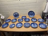 Full lot of 16 blue and white Christmas collector plates arranged on table showing detailed traditional winter scenes.