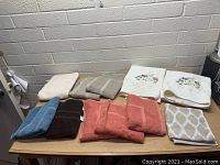Lot of 12 new towels arranged on a table featuring multiple colors and designs, including coral, brown, blue, beige, white with floral embroidery, and patterned towels.