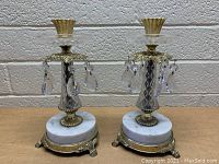 Front view of a pair of vintage candle holders with round white marble bases, gold-tone feet and metal details, crystal prisms, and glass bobeche holders.