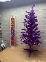 4-foot purple tinsel Christmas tree lit with red lights displayed next to its original cardboard box showing the tree image and branding.