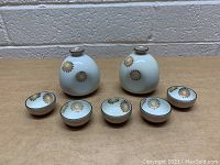 Two porcelain teapots and five matching teacups with yellow and brown floral designs on a white background, displayed on a wooden surface against a brick wall.