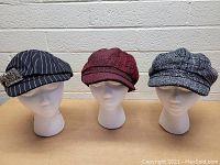 Front view of three women's hats on display stands against a brick wall, showing different patterns and styles.