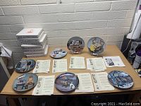 Overview of 7 Bing & Grondahl collector plates arranged on table with certification papers and stacked boxes in background.