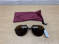 Front view of Suncloud polarized aviator sunglasses with brown lenses and gold metal frames, placed with the maroon protective pouch.