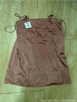 Front view of a large brown dress with thin shoulder straps lying flat on a tiled floor. The dress shows some wrinkles and a tag indicating size 33 is visible.