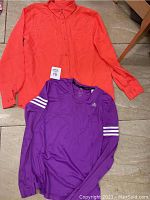 Overview of two small-sized shirts on floor. Orange collared long sleeve shirt and purple Adidas long sleeve athletic shirt with white stripes on sleeves.