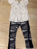 Top-down photo of the outfit laid flat showing white floral patterned shirt and black gray leggings with side panels.