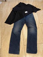 Photo of Guess blue denim jeans size 32 paired with plain black short-sleeve shirt on floor.