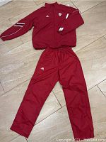 Full view of Adidas red jacket and pants laid flat, showing IU and Adidas logos, sleeve stripe details, size Medium tag visible