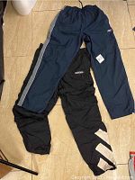Two pairs of sports pants laid flat on floor, one black Adidas with white designs, one navy blue KGR with side stripes
