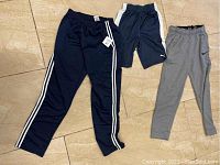 Three pairs of athletic bottoms displayed on floor: navy Adidas track pants, black Puma shorts, gray Nike sweatpants.