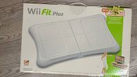 Boxed Wii Fit Plus Balance Board lying on floor, showing front of the box and balance board image.