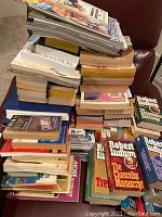 Large pile of assorted paperback books and magazines on a chair and adjacent area.