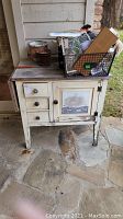 Photo of weathered wooden cabinet with drop side door closed, three small drawers on left, metal knob handles, and various items on top.