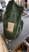 Green storage bag containing disassembled artificial Christmas tree with clear plastic pocket holding assembly instructions.