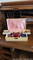 Photo of wood jewelry box with open lid and pink interior lining showing compartments filled with assorted costume jewelry including bracelets and brooches.