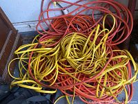 Photo showing a pile of tangled yellow flat and orange round extension cords on a gray surface.