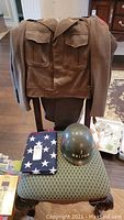 Chair with two WWII uniform jackets hanging, matching trousers below, M1 helmet and folded flag on seat