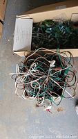 Box containing tangled holiday string lights with green and brown wires and clear bulbs, cords and plugs visible