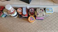 Photo showing an assortment of tins of various shapes and designs arranged on a carpeted floor, including circular, square, and rectangular tins with colorful vintage-style artwork.