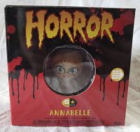 Front view of unopened boxed figure showing horror-themed black and red packaging and Annabelle doll visible through round window.