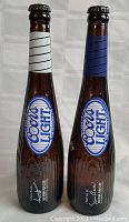 Front view of two Coors Light beer bottles with baseball bat themed designs, one with white/black grip tape and one with blue grip tape, standing upright on a white background.