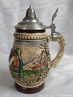 Side view of German beer stein showing detailed relief with mountain, house, and outdoor scene under ornate metal lid.