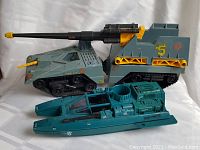 Gray-green armored tank with large cannon and black caterpillar tracks, number 5 and Cobra emblem decals visible, alongside a teal blue hovercraft vehicle with cockpit and engine details.