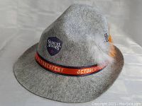 Grey felt Oktoberfest style hat with Samuel Adams logo patch, orange ribbon band, and white/orange feathers attached.
