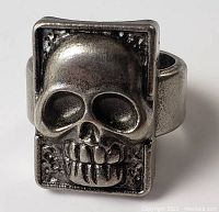 Front view of rectangular silver-tone metal ring with raised skull design and adjustable band.