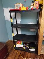4 tier black plastic shelving unit shown empty in corner of room with blue walls, yellow locking tabs visible on support poles, black shelving surfaces clear of items except for few small objects on bottom shelf.