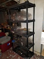 Black plastic shelving unit with multiple shelves, each shelf has grid pattern with items and clear plastic storage trays on it, set against a white wall.