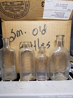Four small antique clear glass bottles displayed in front of a cardboard box labeled 'Sm. Old Bottles'