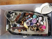 Top view of mixed jewelry and accessories inside a plastic storage container, showing various types of jewelry and a cosmetic item