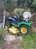 Overview photo of John Deere 325 riding lawn mower from side showing green body, yellow cutting deck, rear and front tires, supervising environment outdoors.