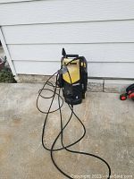 Full view of Karcher power washer showing body, attached power cord, and hose on concrete floor against siding.