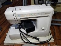 Full view of Kenmore sewing machine with power cord and foot pedal wrapped around its base, showing compact cream colored unit in plastic carrying case.