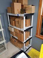 Plastic shelving unit with four shelves, white shelves, black supports, boxes stored on unit, near window.