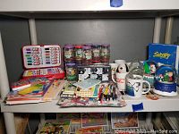 Full view of various Snoopy and Peanuts collectibles including mugs, jars, stationery, pens, a stuffed toy, and snow globes arranged on shelves.